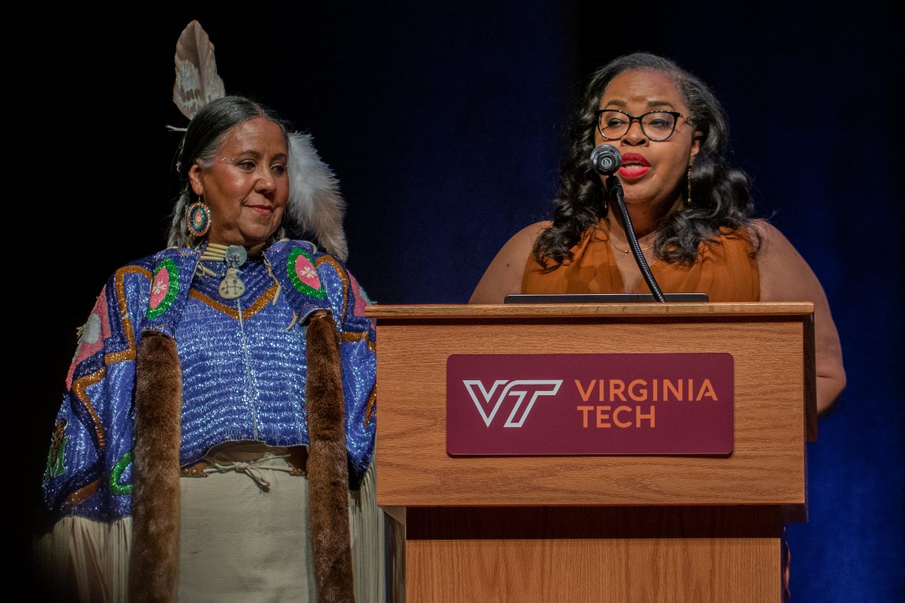 Two women are on stage, one stands behind a podium, speaking into a microphone. The other one stands to her side, attired in a traditional Native American ensemble.