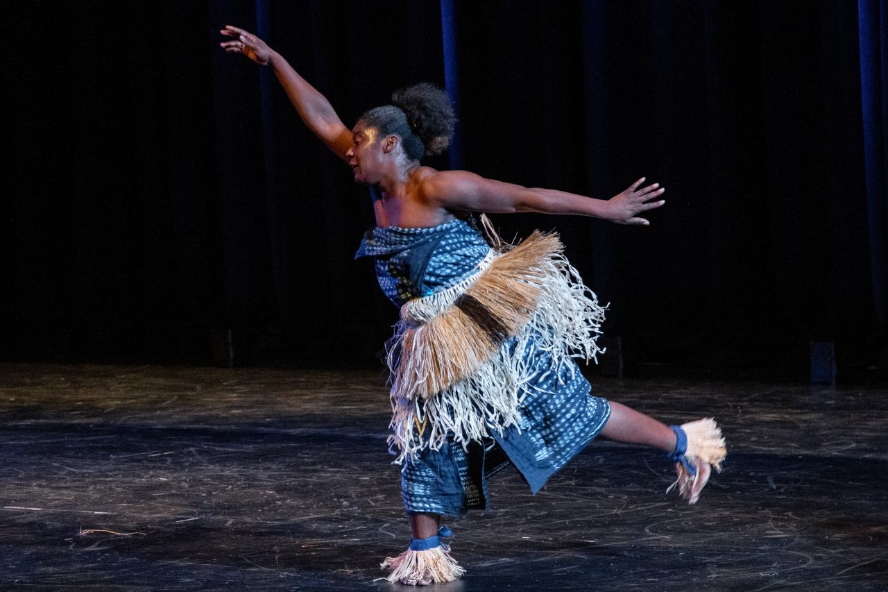 A woman dances across a stage with arms outstretched as she moves. She is wearing blue print with a grass skirt around her waist and grass also circles her ankles.