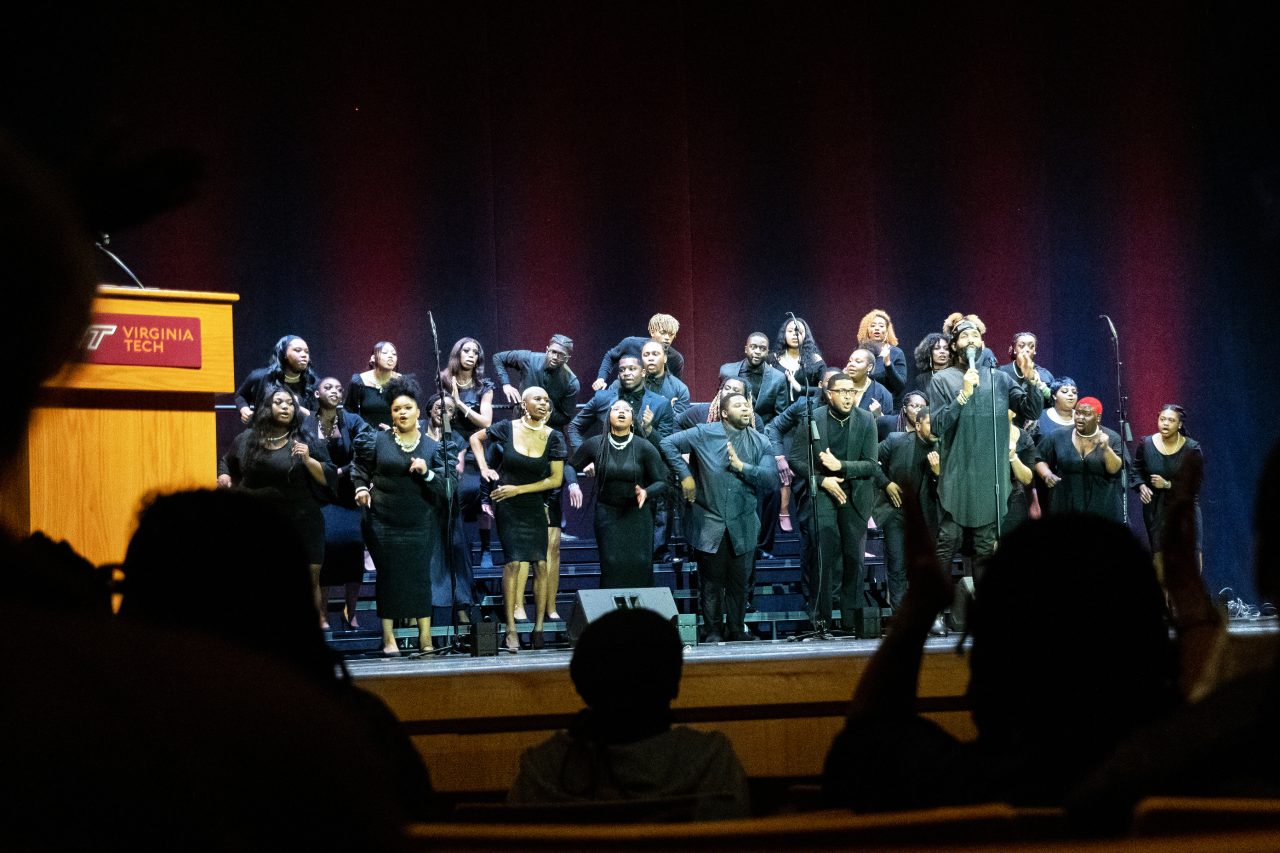 A large group of people are arranged on risers on a stage. They are singing and clapping to the music. 