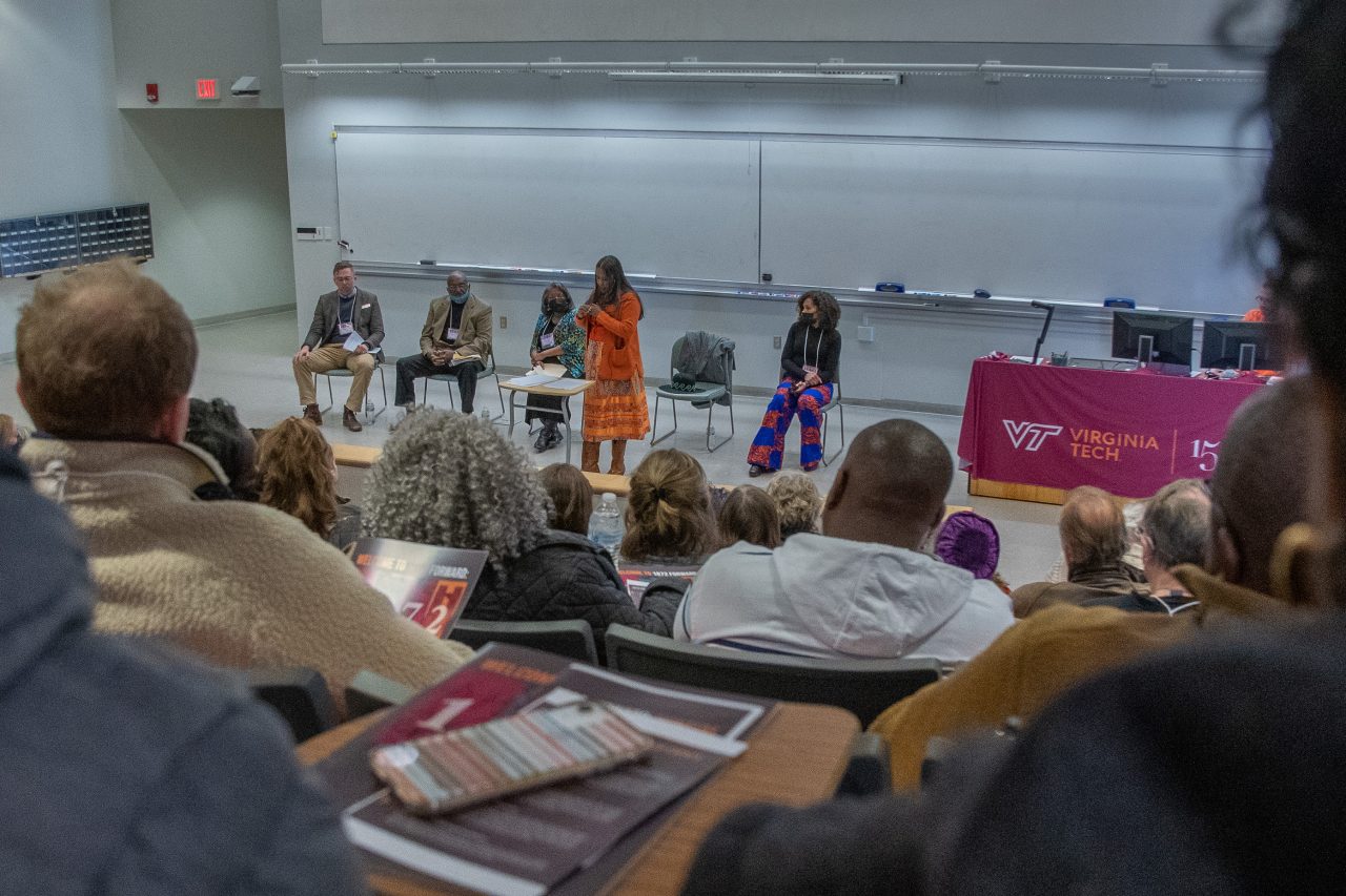 Looking through a crowd from the back of a lecture style classroom, four people are seated across the front of the room. Another person wearing an orange skirt and top is standing while speaking to the attendees. 