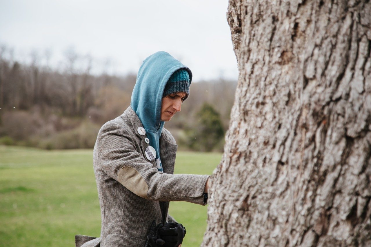 A person wearing a hooded sweatshirt touches the trunk of a tree with their head bowed. 