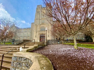 A gray Hokie Stone Burruss Hall on a sunny day while snow sprinkles onto green grass.