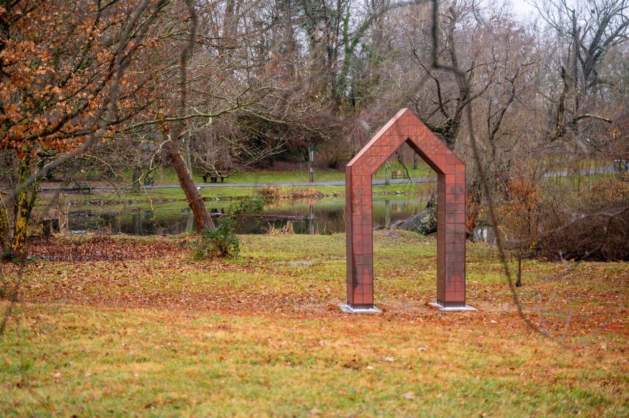 A tiled archway sits in the distance on the lawn. In the background is the edge of a pond, a pathway, and trees. 
