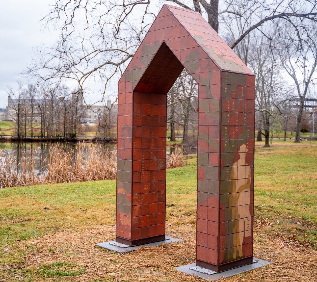 A tiled archway sits on a lawn. To the left of the arch is a pond and trees appear in the background. On the archway are depicted historical imagery of Virginia Tech. 